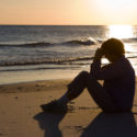 Mature woman sits on the beach with her head bowed and praying as the sun sets on the water.